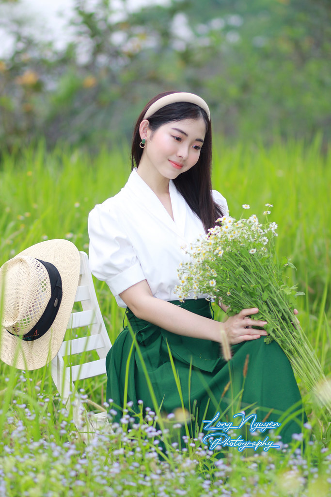 A beautiful girl donning a white shirt, a green waist-belted skirt, and a turban and holding white daisies graces the green lawn with some wildflowers and the chair, her dark hair subtly mixed with red.